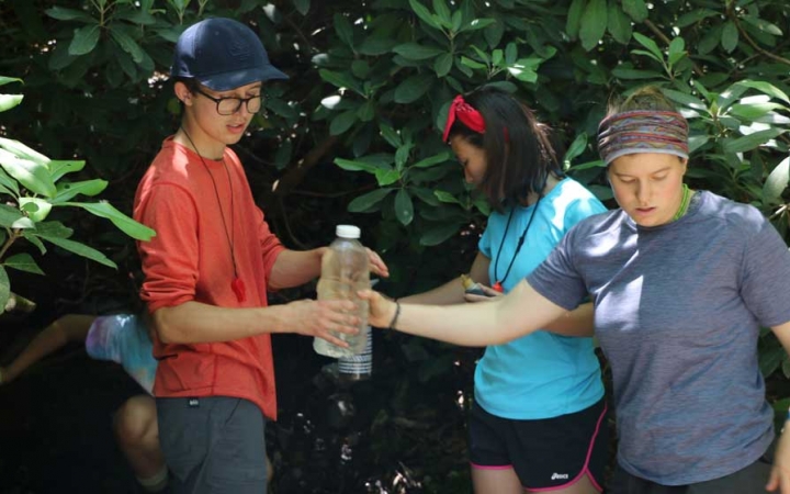 Two students pass a bottle of water between them. There are other students and green leaves in the background. 